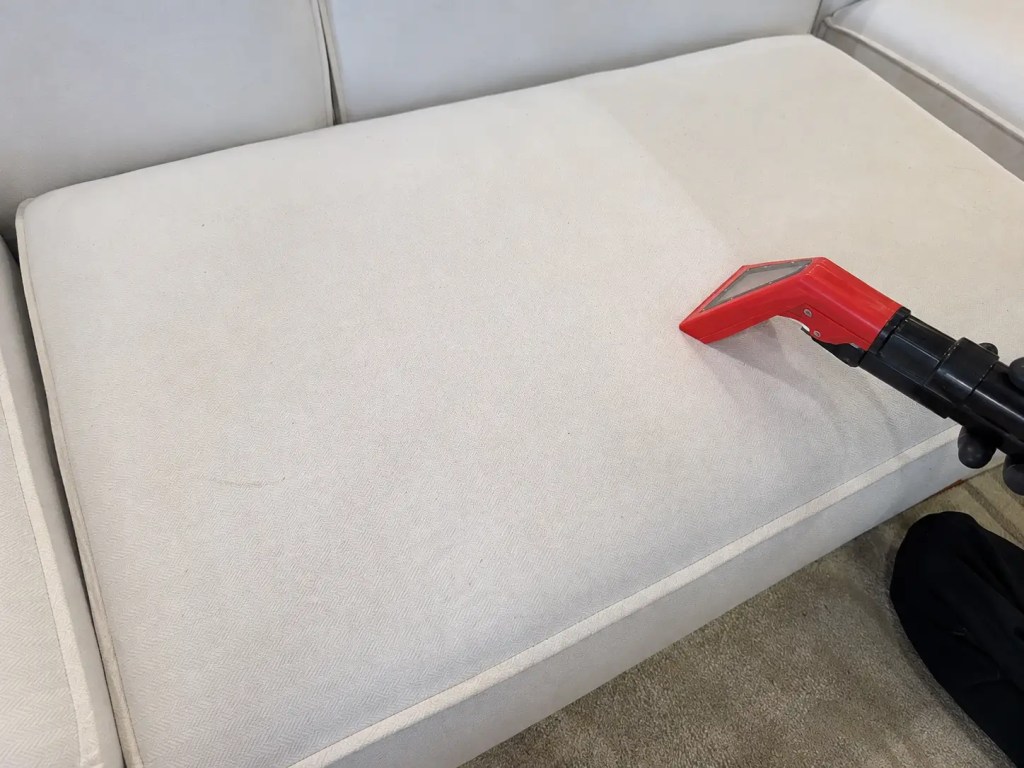 Red upholstery extraction tool cleaning a light-colored couch cushion